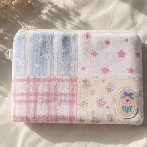 May include: A rectangular zippered pouch featuring a patchwork design of four distinct fabric squares. The patterns include light blue with white stars, white with pink stars, pink plaid, and cream with small pink flowers, plus a cupcake patch.