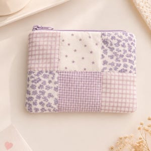 May include: A small, rectangular, patchwork-style zippered pouch in shades of lavender and white. The design features squares with floral, star, and gingham patterns. The zipper and stitching are lavender. The pouch is likely for carrying small items.