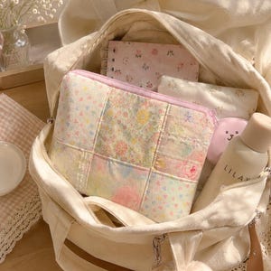 Puede incluir: Un bolso de lona color crema que contiene un estuche con cremallera de patchwork con estampado floral, un cuaderno de espiral con cubierta de flores rosas, una botella peque&ntilde;a beige con la etiqueta LILAY y un llavero de peluche con lazo rosa.