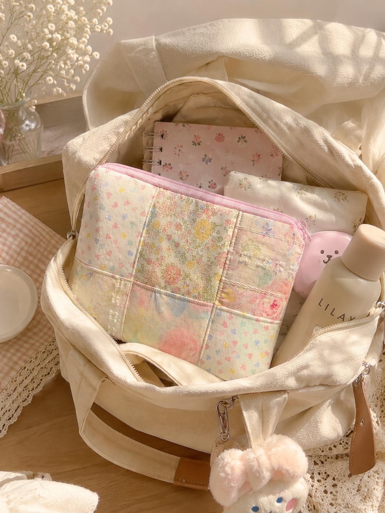 Puede incluir: Un bolso de lona color crema que contiene un estuche con cremallera de patchwork con estampado floral, un cuaderno de espiral con cubierta de flores rosas, una botella peque&ntilde;a beige con la etiqueta LILAY y un llavero de peluche con lazo rosa.