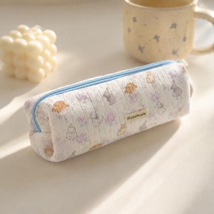 May include: A cylindrical pencil case made of white fabric with a subtle vertical ribbed texture, featuring a repeating pattern of small brown and gray rabbits and purple floral accents. It has a light blue zipper and a small beige tag labeled Handmade.