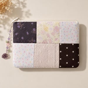 May include: A rectangular zippered pouch featuring a patchwork design of six fabric panels. The panels include black floral, yellow floral, pastel floral, beige textured lace, and black with white stars. A beaded charm with a grape cluster hangs.