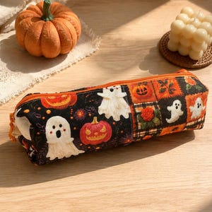 May include: A rectangular fabric pencil case featuring a black background with a repeating pattern of white ghosts, orange jack-o'-lanterns, and small floral motifs. The case has an orange zipper closure along the top edge and a structured shape.