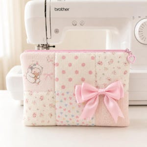 Puede incluir: Un estuche de tela rectangular con un diseño de patchwork en tonos rosa y crema. El patrón incluye lunares, pequeños estampados florales y una ilustración de una oveja. Un gran lazo de satén rosa está sujeto en la parte frontal.