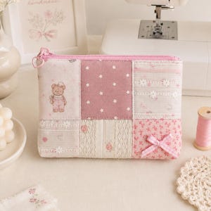 May include: A rectangular fabric zipper pouch featuring a patchwork design of pink and white textiles. The pattern includes a teddy bear illustration, white stars on a dusty rose background, lace trim, floral prints, and a small pink gingham bow.
