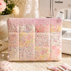 Peut inclure: Une pochette rectangulaire zippée en tissu matelassé avec un motif patchwork composé de divers imprimés floraux dans des tons rose, jaune et blanc. Le bord supérieur est orné d'une dentelle rose et d'une tirette de fermeture en forme de cœur.