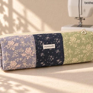 May include: A rectangular fabric pencil case featuring a patchwork design of floral patterns in shades of lavender, navy blue, and sage green. A small rectangular label with the word Handmade is stitched onto the centre panel. It has a zip closure.