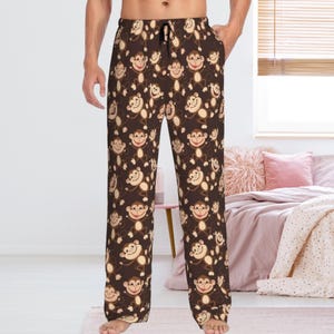 May include: Dark brown lounge pants featuring an all-over repeating pattern of light brown cartoon monkeys with smiling faces. The pants have a black drawstring at the elasticized waistband and a straight-leg fit, providing a casual look.