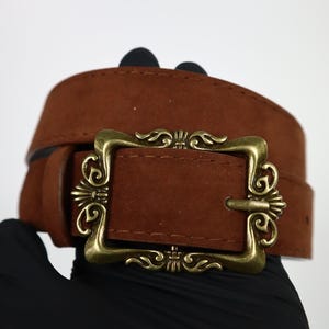 Könnte beinhalten: Ein brauner Wildleder-Gürtel mit einer dekorativen goldfarbenen Schnalle. Die Schnalle hat ein verziertes Design mit Wirbelmustern. Der Gürtel hat eine mittelbraune Farbe und scheint aus Wildleder zu sein.