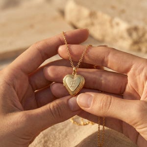 May include: A gold-colored heart-shaped locket necklace with a delicate chain. The locket features a beaded border and an engraved letter "E" in the center. The necklace is held by a person's hands.