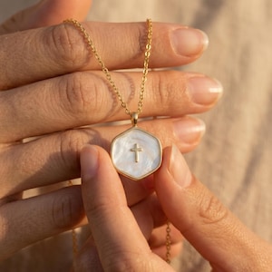 Pode incluir: Colar dourado com um pingente hexagonal. O pingente tem um centro branco perolado e uma pequena cruz dourada. A corrente é delicada. O colar é segurado pelas mãos de uma pessoa. Símbolo religioso.