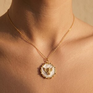 May include: Gold-toned necklace featuring a circular pendant with a gold angel design on a white background. The pendant is framed by a gold border. The necklace is worn around the neck.