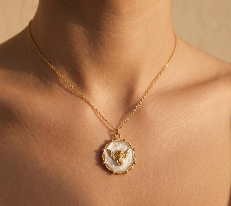 May include: Gold-toned necklace featuring a circular pendant with a gold angel design on a white background. The pendant is framed by a gold border. The necklace is worn around the neck.