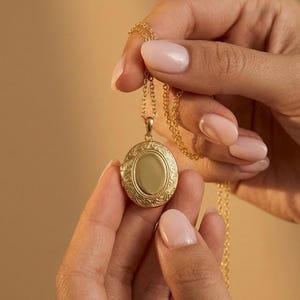 以下が含まれることがあります： 金色のロケットネックレス。楕円形のペンダントには、縁に装飾的な彫刻が施され、中央は滑らかです。ネックレスは人が持っています。