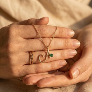 May include: A gold-colored necklace with the letter "A" pendant, featuring a small green gemstone. The necklace is held in a person's hands, along with a gold ring. The jewelry is set against a neutral background.