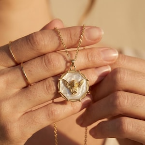 May include: Gold-toned necklace featuring a pendant with an angel design. The pendant has a white background with a gold angel figure in the center. The necklace is held by a person.