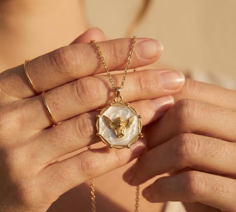 May include: Gold-toned necklace featuring a pendant with an angel design. The pendant has a white background with a gold angel figure in the center. The necklace is held by a person.