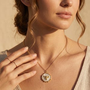 May include: A gold-toned necklace with a pendant featuring a white background and a gold-colored winged design. The necklace is worn around the neck. The image is a close-up shot of the necklace and the person wearing it.