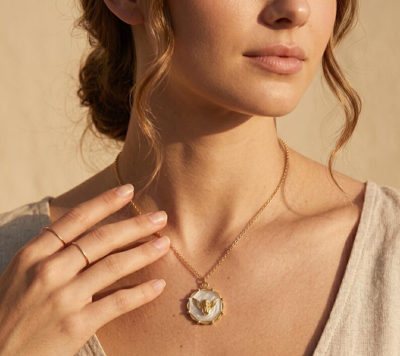 May include: A gold-toned necklace with a pendant featuring a white background and a gold-colored winged design. The necklace is worn around the neck. The image is a close-up shot of the necklace and the person wearing it.