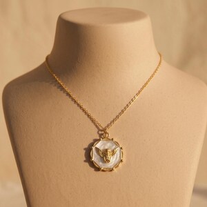 May include: A gold-colored necklace with a pendant featuring a winged figure. The pendant has a white background and a gold-colored frame. The necklace is displayed on a mannequin.