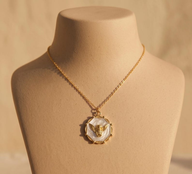May include: A gold-colored necklace with a pendant featuring a winged figure. The pendant has a white background and a gold-colored frame. The necklace is displayed on a mannequin.