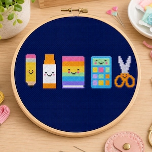 Può includere: Un telaio da ricamo circolare in legno contenente un tessuto blu scuro con un disegno a punto croce di cinque articoli scolastici: una matita gialla, una colla stick bianca, un libro arcobaleno, una calcolatrice blu e forbici arancioni.