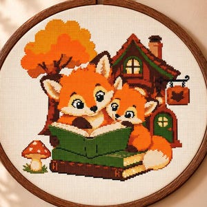 May include: A circular wooden embroidery hoop containing a cross-stitch pattern of two orange foxes reading a green book in front of a brown house and an orange tree. A small red and white mushroom is stitched to the left of the foxes.