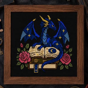 May include: A framed cross-stitch embroidery featuring a blue dragon with yellow horns perched atop a stack of open books, flanked by pink roses on a black background. The dragon has blue wings with yellow star accents and a crescent moon on the book.