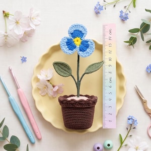 Puede incluir: Una flor de ganchillo azul y amarilla en una maceta marrón, sobre un plato amarillo. Una regla muestra medidas de hasta 14 cm. También se ven ganchillos y tijeras, junto con otras flores.