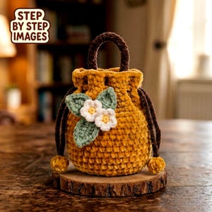 Puede incluir: Un bolso de ganchillo amarillo mostaza hecho a mano con un asa y un cordón marrón. El bolso está decorado con una aplicación de hoja verde y flor blanca. El texto "STEP BY STEP IMAGES" está en la esquina superior izquierda.