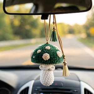 May include: A green and white crochet mushroom car charm hanging from a car's rearview mirror. The mushroom has a green cap with white spots and a white stem. The charm is adorned with tassels and gold accents.