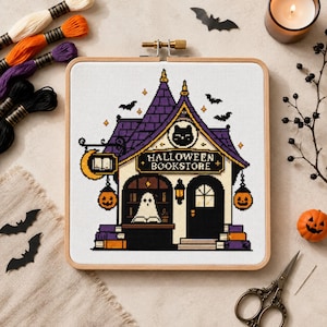 Puede incluir: Un bastidor de bordado cuadrado de madera que contiene un patrón de punto de cruz de una librería de Halloween. El diseño presenta un edificio con techo morado, un fantasma, un emblema de gato negro y el texto HALLOWEEN BOOKSTORE, con calabazas.