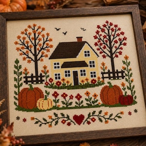 May include: A framed cross-stitch embroidery featuring a yellow house with a dark roof, surrounded by autumn trees, orange pumpkins, and red flowers. The design includes a central red heart at the bottom and small black birds flying in the sky.