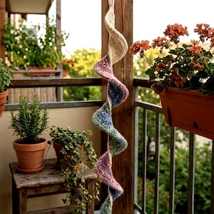 Op de afbeelding: Een handgemaakte, spiraalvormige gehaakte hangdecoratie. De spiraal heeft afwisselende secties van crème, lavendel, blauw en groen garen. Het decoratieve stuk hangt aan een houten balk, met potplanten en een balkonreling op de achtergrond.