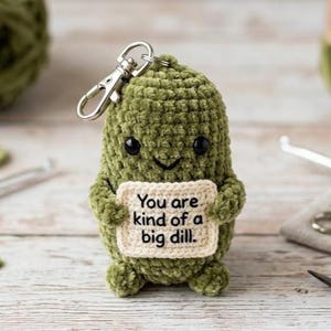 May include: A handmade green crochet pickle keychain with a silver clasp. The pickle has black eyes and a smiling mouth, holding a cream-colored sign that reads "You are kind of a big dill."