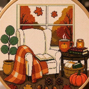 May include: A cross-stitch embroidery in a wooden hoop depicting a white armchair with an orange plaid blanket and an open book. Beside it, a side table holds a pumpkin mug, books, and a candle, with a potted plant and pumpkin nearby.