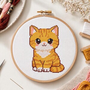 May include: A cross-stitch embroidery of a cartoon orange tabby cat with large black eyes, pink ears, and a white chest. The cat is centered in a wooden embroidery hoop. Embroidery floss and other craft supplies are in the background.