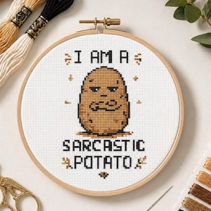 May include: A cross-stitch design in a wooden hoop shows a brown potato with a grumpy face. The text reads "I AM A SARCASTIC POTATO" in black thread. Embroidery threads and gold scissors are also visible in the image.