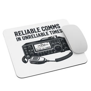 Puede incluir: Alfombrilla de ratón blanca con el texto "RELIABLE COMMS IN UNRELIABLE TIMES" y una ilustración detallada de un transceptor de radio. La radio tiene una pantalla que muestra "14.250 MHz" y el modelo "ICOM IC-7900". Un ratón de ordenador blanco está al lado.