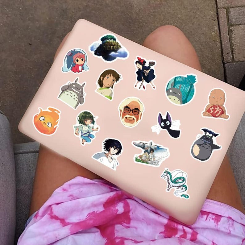 May include: A collection of colorful anime-themed stickers, featuring characters from various animated films, are arranged on a light pink laptop. The stickers include images of characters, creatures, and scenes from popular Japanese animated movies.