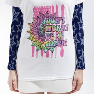 May include: A white short-sleeved t-shirt with long dark blue sleeves featuring a light blue star pattern. The front displays a tie-dye sunflower, a pink distressed American flag graphic, and the text Don't Worry Be Hippie with a peace sign.