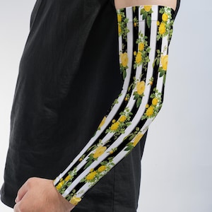 May include: An arm sleeve featuring a pattern of vertical black and white stripes overlaid with clusters of yellow roses and green leaves. The sleeve is worn on the forearm and extends toward the elbow, contrasting with a plain black t-shirt.