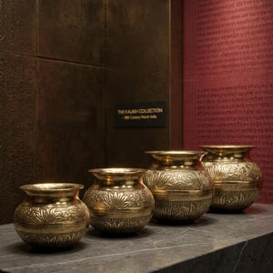 Puede incluir: Cuatro ollas ornamentadas de metal dorado con patrones florales y geométricos. Las ollas están dispuestas en fila sobre una superficie gris oscuro. El texto en la pared dice "THE KALASH COLLECTION - 18th Century North India."