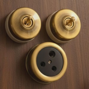 May include: Two circular brass toggle light switches with white bases and one circular brass electrical outlet with a black center. Each component features a metallic finish and a round design, mounted as a set of three individual electrical fixtures.