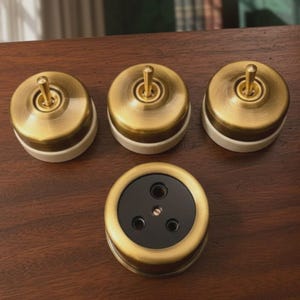 Op de afbeelding: Drie ronde lichtschakelaars met koepels van geborsteld messing en witte keramische voetstukken, elk voorzien van een centrale messing tuimelschakelaar. Daaronder bevindt zich een rond stopcontact met een rand van geborsteld messing en zwart.