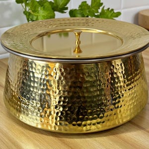 May include: A gold-colored, hammered metal serving bowl with a matching lid. The bowl has a rounded shape with a textured surface and a silver-colored rim. The lid features a small, decorative handle.