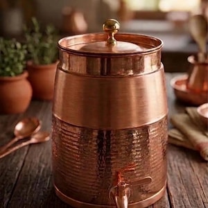 May include: A copper water dispenser with a hammered texture on the lower half and a smooth band in the middle. It has a spigot and a lid with a small, round handle. The dispenser is sitting on a wooden surface.