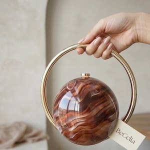 Peut inclure: Un sac à main sphérique avec un motif tourbillonnant marron et ambre, tenu par une poignée circulaire dorée. Le sac a un petit dessus doré et une étiquette qui dit "BeCelia".