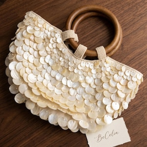 Pode incluir: Uma bolsa de mão semicircular coberta com discos de madrepérola cor de creme sobrepostos. A bolsa possui duas alças redondas de madeira castanho-escura presas com alças de tecido bege. Uma pequena etiqueta branca com o texto BeCelia está presa à bolsa.