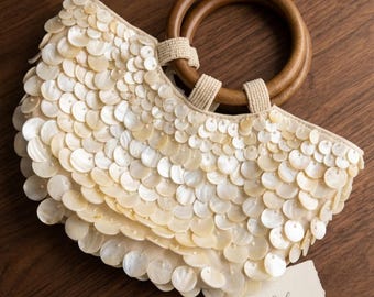 Puede incluir: Un bolso de mano semicircular cubierto con discos de nácar de color crema superpuestos. El bolso cuenta con dos asas redondas de madera marrón oscuro unidas con lazos de tela beige. Una pequeña etiqueta blanca con el texto BeCelia está unida al bolso.